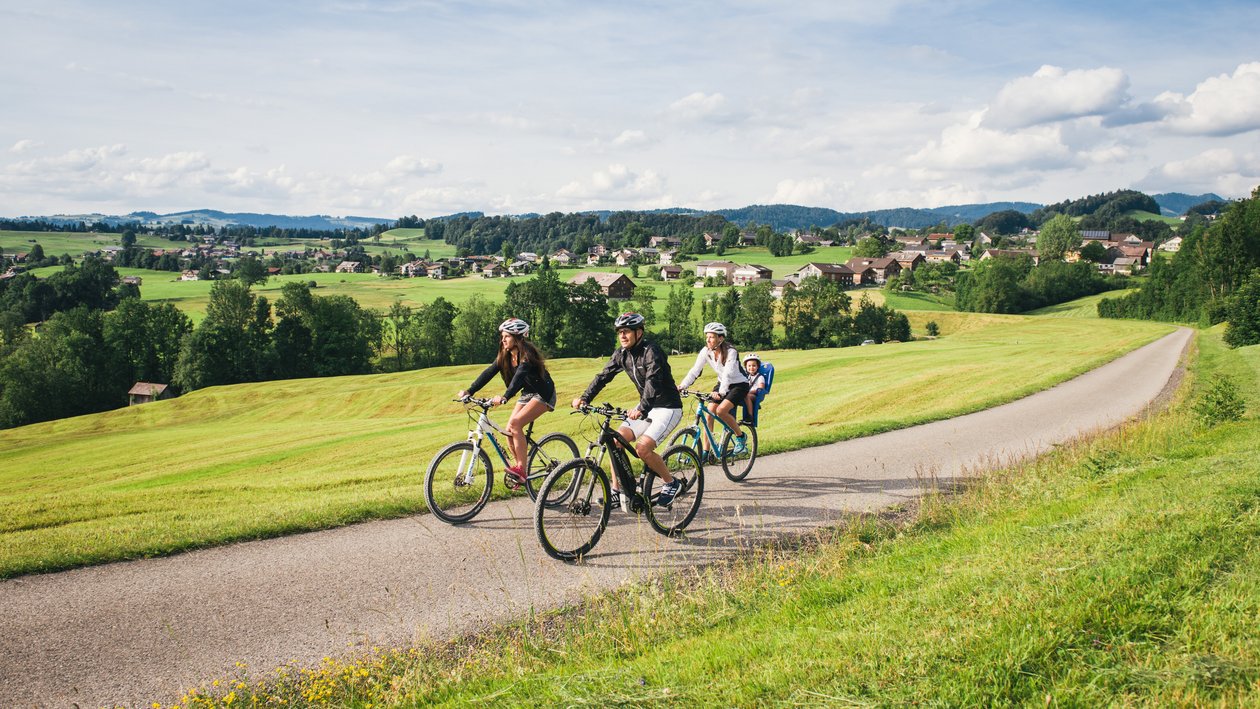 Radfahren im Bregenzerwald Drei Radfahrer auf einem befestigten Weg in einer grünen Landschaft mit Hügeln und einem Dorf im Hintergrund.