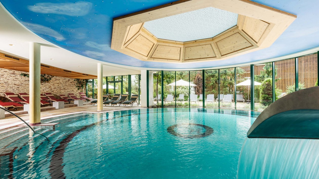 Wohlfühlmomente im hirschen spa Innenansicht des Hotelspas mit Pool und Wasserfall, umgeben von Liegen und Fenstern